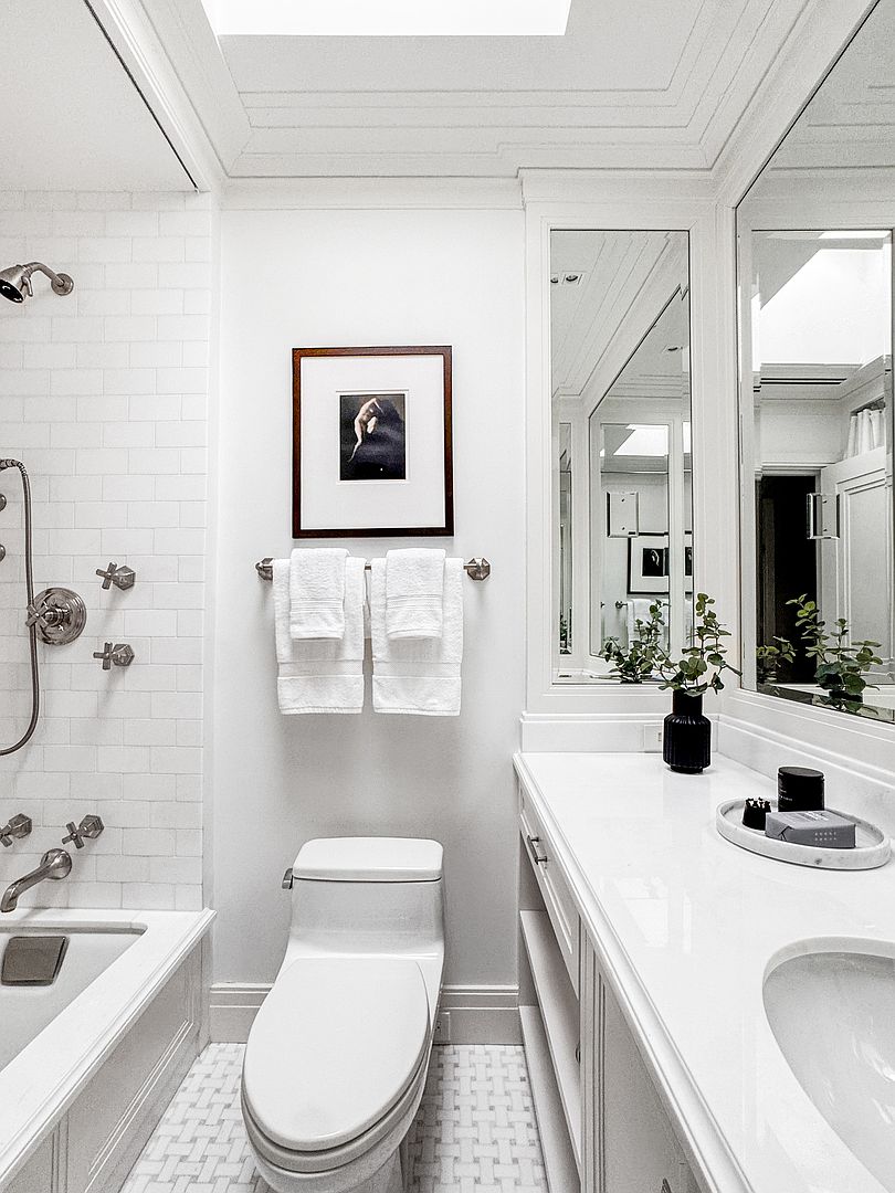 This bright, modern bathroom features a clean white aesthetic with a subway-tiled shower surround and a classic basketweave-patterned floor. The space includes a sleek vanity with a white countertop, a wall-mounted toilet, and a framed piece of art above the towel rack. A large mirror reflects the room, enhancing the sense of light and space in this well-appointed bathroom.