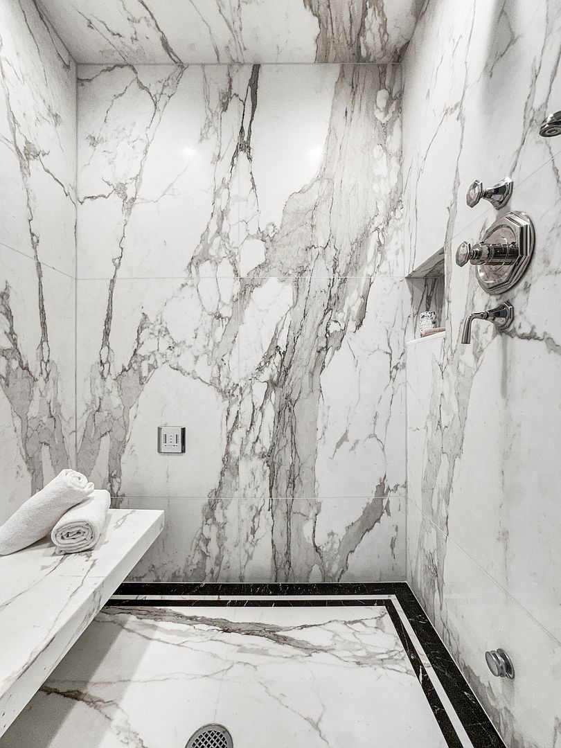 This luxurious walk-in shower features expansive, light-colored stone slabs with dramatic veining covering the walls and bench. A contrasting dark stone border frames the floor, which is finished in matching light stone, while polished chrome fixtures and a built-in niche add a refined touch. The perspective is a low-angle, eye-level shot that emphasizes the height and high-end materials of the shower enclosure.