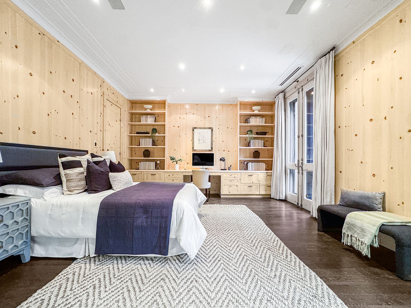 This spacious primary bedroom features light wood paneling on the walls and a built-in desk and shelving unit that spans the back wall. A large bed with dark bedding sits in the foreground, complemented by a patterned area rug and a bench at the foot of the bed. Tall glass doors with light curtains provide natural light and access to the outdoors, creating a warm and sophisticated atmosphere.
