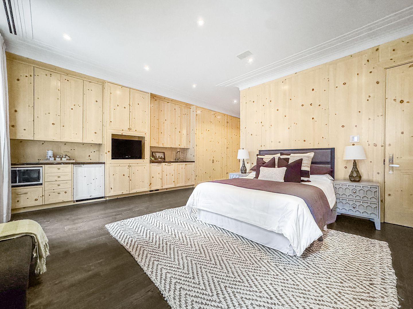 This spacious bedroom features light wood paneling throughout, creating a warm and rustic aesthetic. The room includes a built-in kitchenette area with cabinetry, a microwave, and a small refrigerator, alongside a comfortable bed dressed in neutral linens. A textured, patterned area rug sits on the dark wood flooring, and the space is illuminated by recessed lighting and bedside lamps.