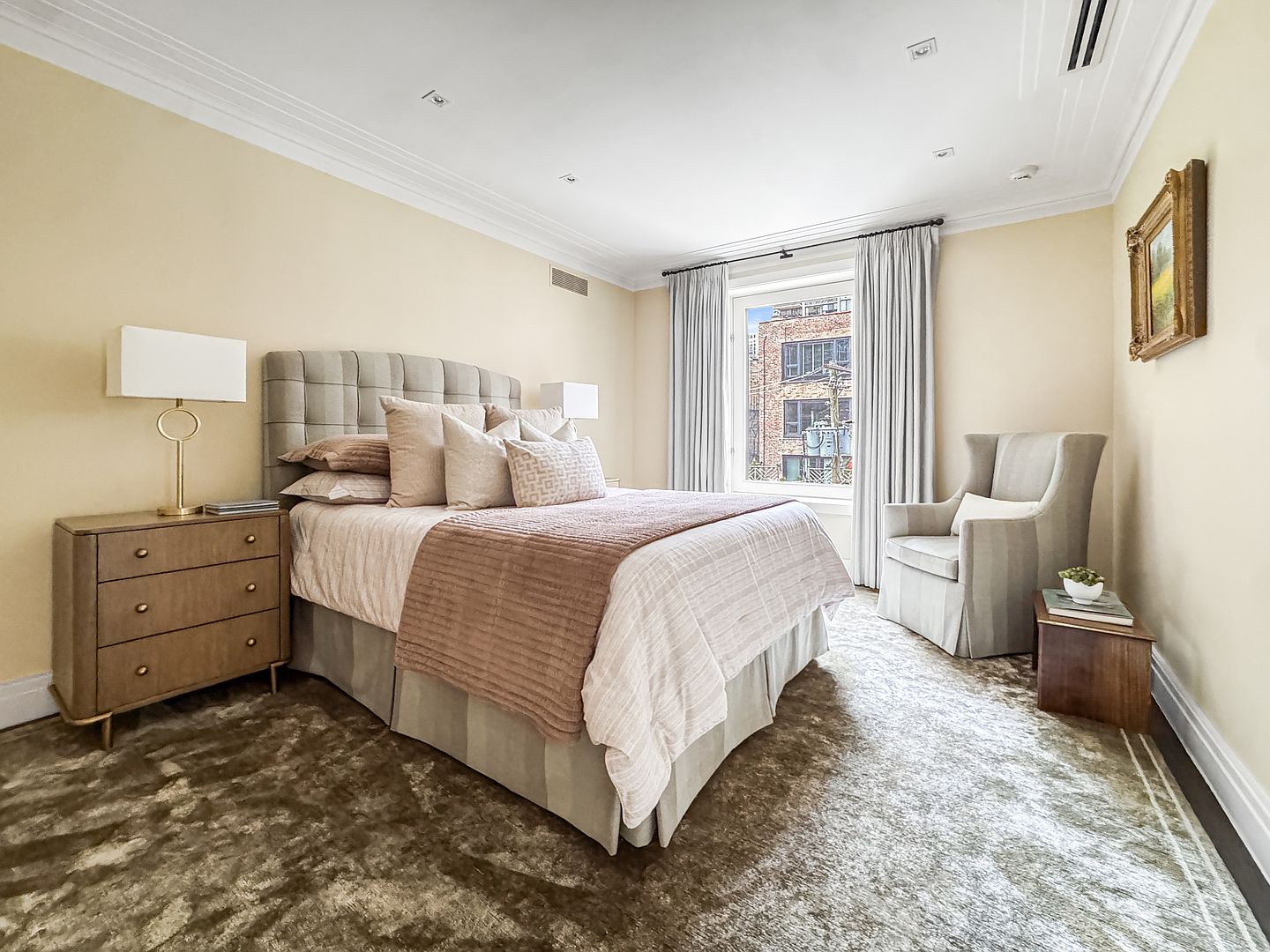 This elegant primary bedroom features a plush, tufted headboard and a neutral color palette that creates a serene and sophisticated atmosphere. A large window framed by floor-length curtains allows natural light to fill the space, while a textured area rug adds warmth to the room. The layout includes a bedside chest, a comfortable armchair in the corner, and a framed piece of art, offering a refined and inviting living space.