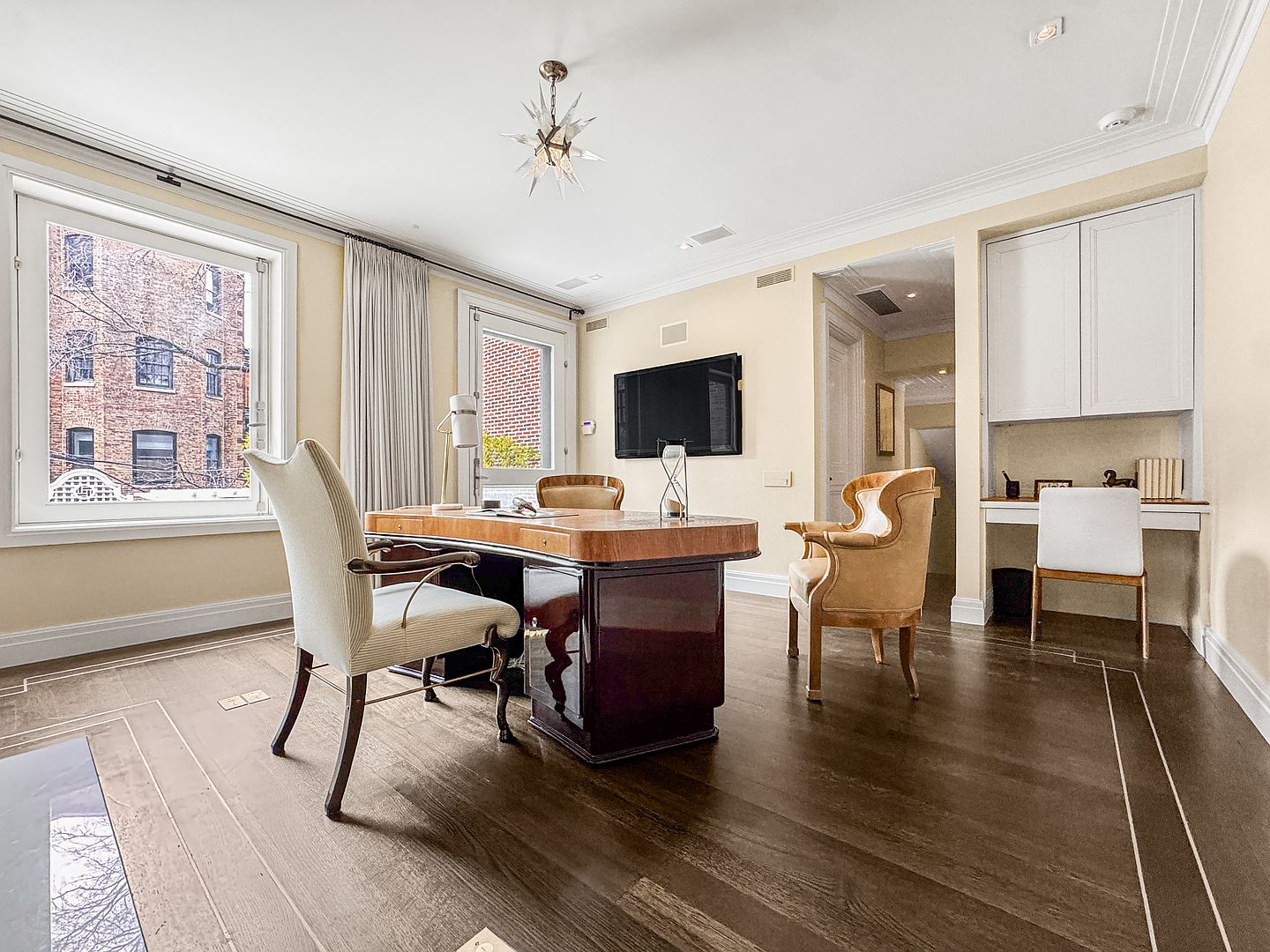 This sophisticated home office features dark hardwood flooring with a decorative inlay border and light-toned walls. The room is anchored by a substantial, polished wood desk and a pair of elegant chairs, while a built-in desk nook with overhead cabinetry provides additional workspace. A large window offers natural light and a view of the exterior, complemented by a modern starburst-style chandelier and a wall-mounted television.