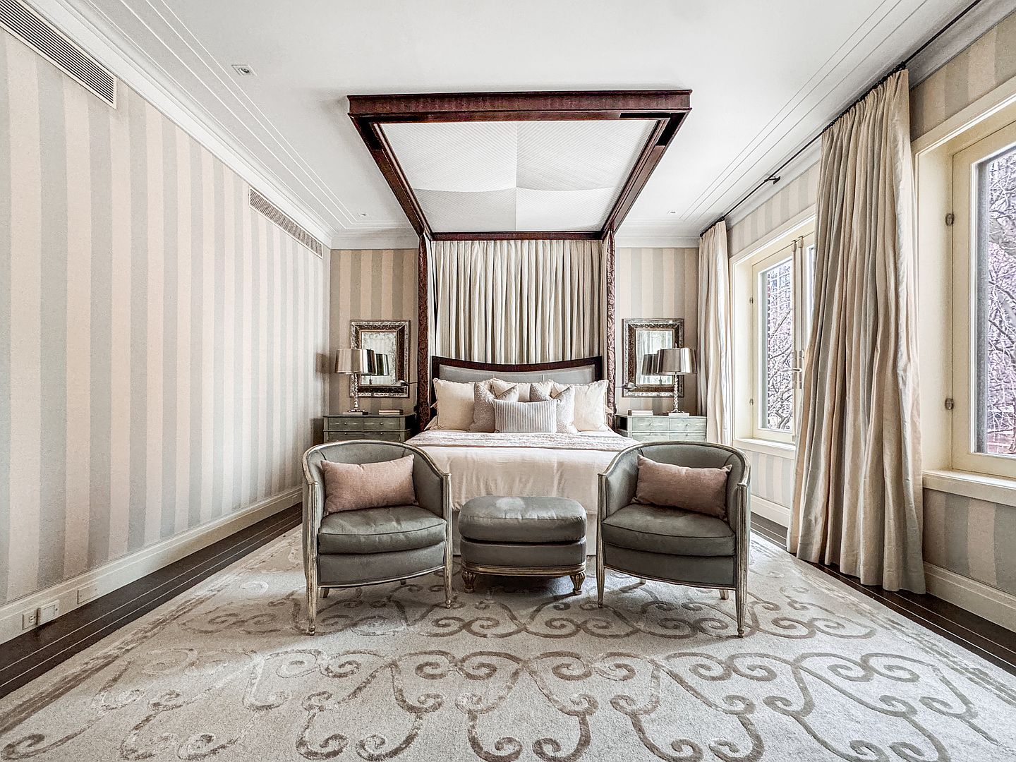 This elegant primary bedroom features a grand four-poster bed as the central focal point, framed by a dark wood canopy and soft, draped fabric. Two matching armchairs and a central ottoman sit atop a large, intricately patterned area rug, creating a sophisticated seating area. The room is finished with striped wallpaper, tall windows dressed in neutral curtains, and symmetrical bedside tables with lamps, offering a refined and tranquil atmosphere.