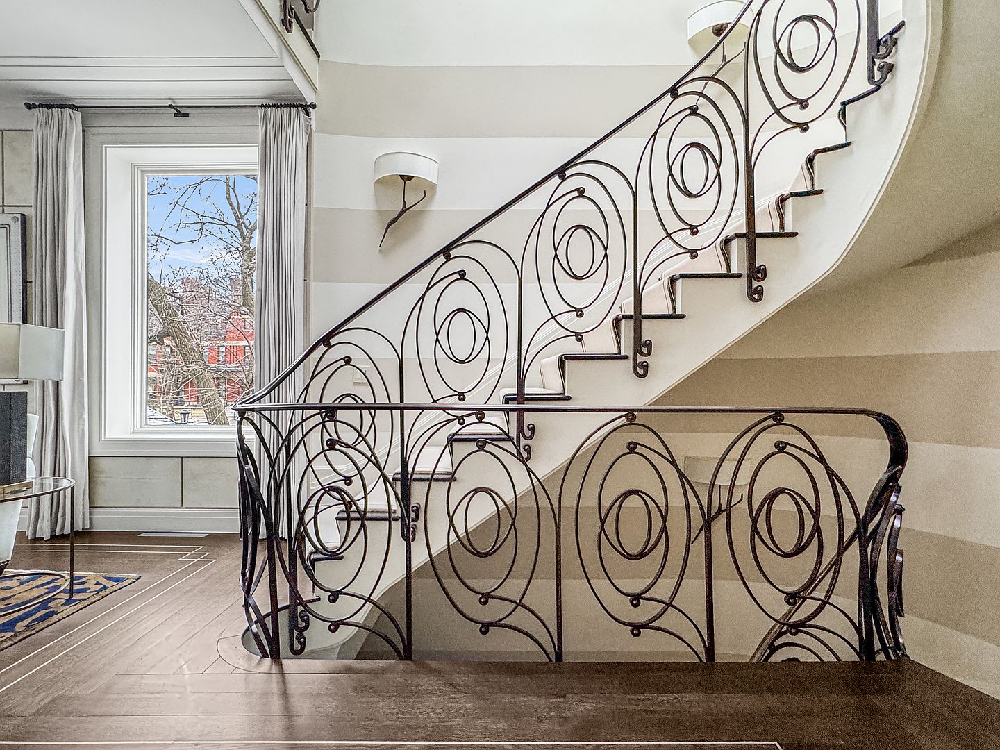 This elegant interior space features a grand, curved staircase with intricate, dark metal railings showcasing a repeating oval design. The walls are adorned with subtle, horizontal striped wallpaper, and a wall-mounted sconce provides soft accent lighting. A large window with light-colored curtains allows natural light to fill the area, highlighting the polished wood flooring and the sophisticated architectural character of the home.