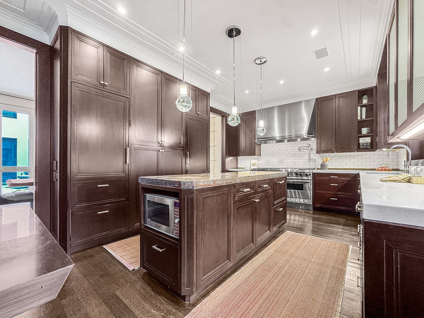This sophisticated kitchen features dark wood cabinetry, a large central island with a stone countertop, and stainless steel appliances, including a professional-grade range. Three glass pendant lights hang above the island, providing focused illumination, while a white subway tile backsplash adds a classic touch. The perspective captures the depth of the room, highlighting the contrast between the dark cabinetry and the light-colored countertops and flooring.