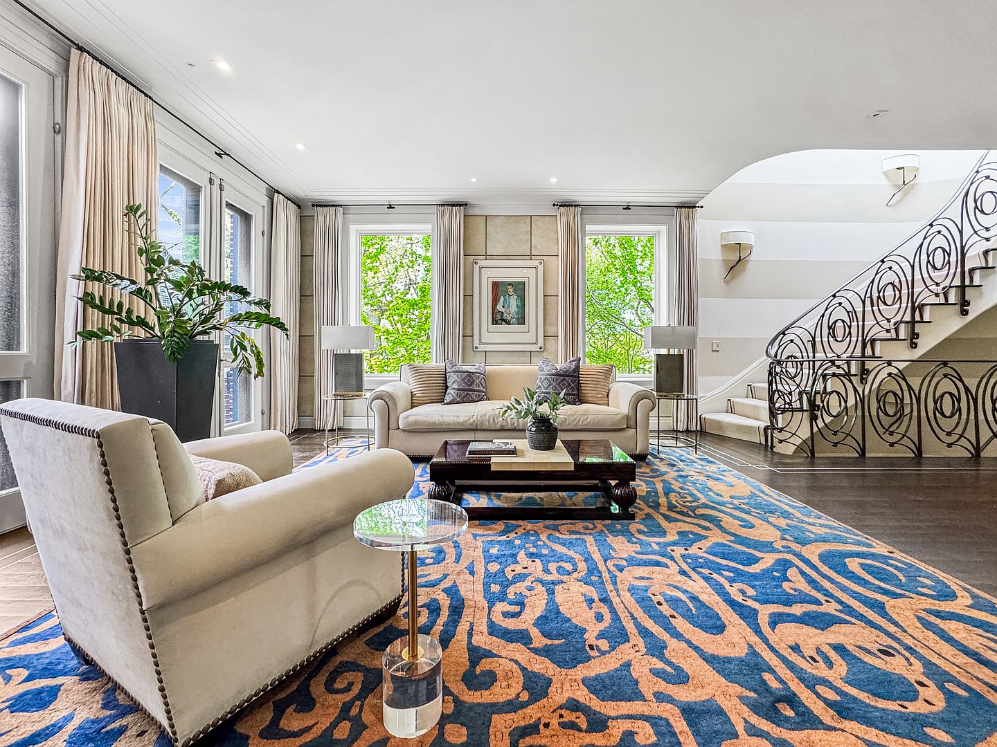 This elegant living room features a sophisticated design with a prominent blue and orange patterned area rug as the focal point. The space is furnished with a plush cream-colored sofa and a matching armchair, complemented by a dark wood coffee table and a sleek glass side table. A grand staircase with intricate wrought-iron railings curves gracefully along the right side, while large windows draped in neutral curtains allow ample natural light to fill the room.