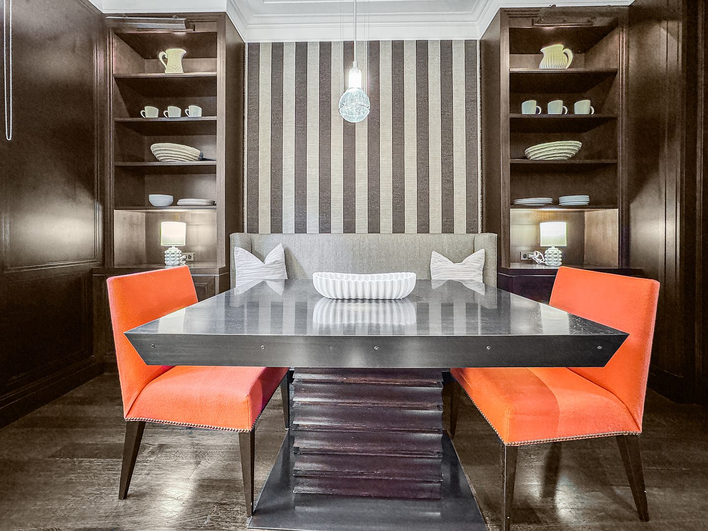 This sophisticated dining area features a dark, tiered-base table flanked by two vibrant orange upholstered chairs and a built-in banquette. The space is framed by symmetrical dark wood shelving units on either side of a striped accent wall, creating a formal and intimate atmosphere. A single glass pendant light hangs above the table, which is centered by a decorative white bowl, while small lamps on the shelves provide warm, ambient lighting.