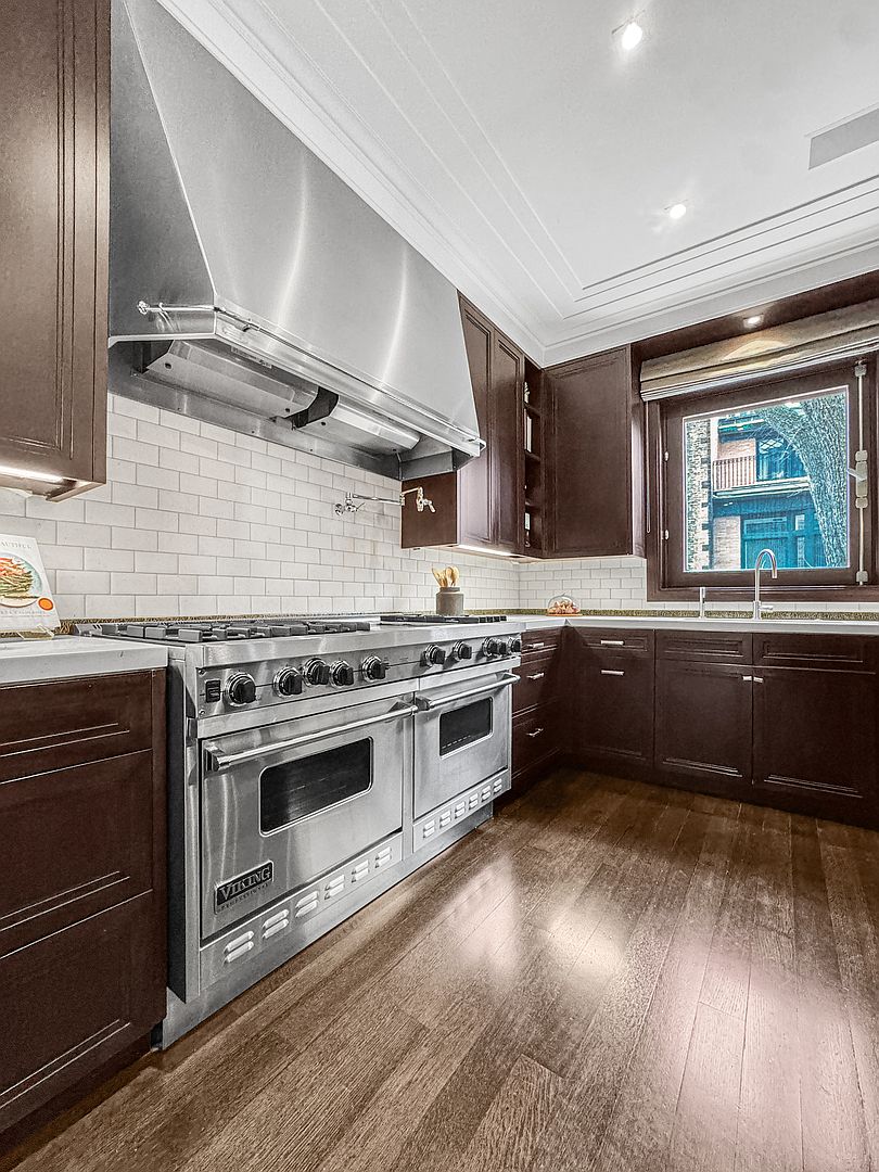 This kitchen features dark wood cabinetry paired with a white subway tile backsplash and a large stainless steel professional-grade range. A prominent stainless steel vent hood sits above the stove, while a window above the sink provides natural light and a view of the exterior. The space is finished with warm-toned wood flooring and recessed ceiling lighting, creating a sophisticated and functional culinary environment.