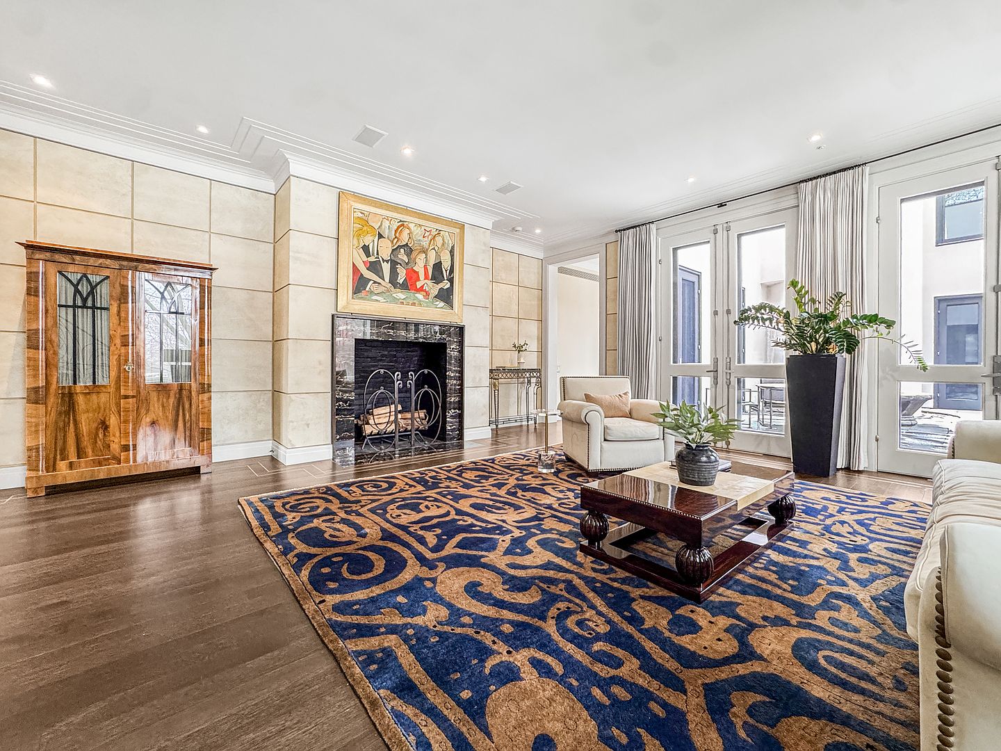 This elegant living room features dark wood flooring topped with a large, ornate blue and gold patterned area rug. A central fireplace with a dark stone surround serves as a focal point, topped by a vibrant framed painting, while a tall wooden cabinet stands to the left. Large glass doors with white frames allow natural light to fill the space, leading out to an outdoor area with a large potted plant nearby.