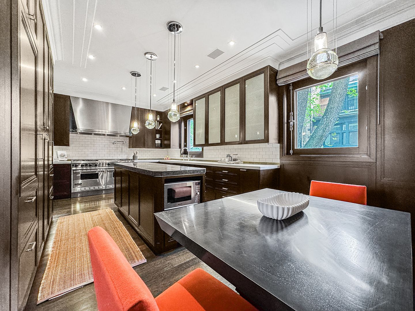 This sophisticated kitchen features dark wood cabinetry, a central island with a built-in microwave, and a professional-grade stainless steel range with a matching hood. A dining area with vibrant orange chairs sits in the foreground, while a large window provides a view of the outdoors. The space is illuminated by modern pendant lighting and recessed ceiling lights, creating a warm and polished atmosphere.