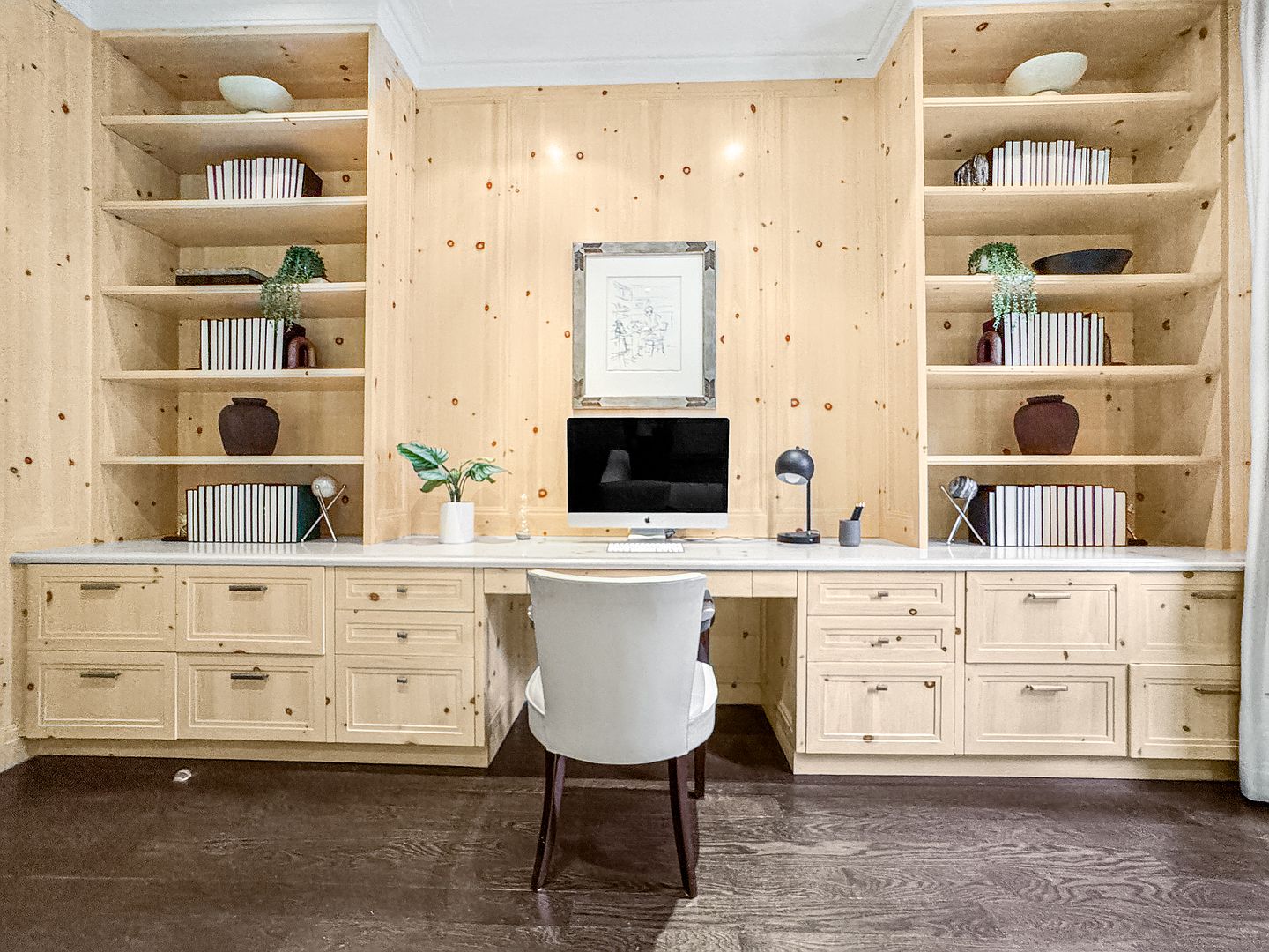This sophisticated home office features extensive custom light wood cabinetry with integrated shelving flanking a central desk area. A sleek computer monitor sits on a light-colored stone countertop, complemented by a modern upholstered chair and minimalist desk accessories. The symmetrical design and warm wood tones create a professional yet inviting workspace.