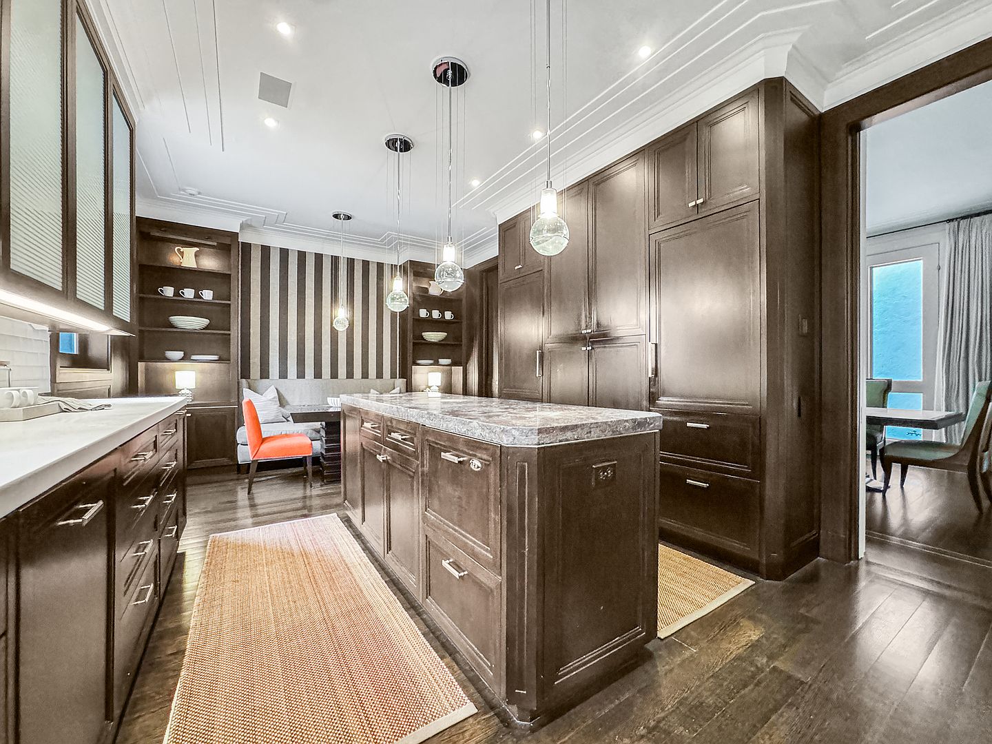 This sophisticated kitchen features dark wood cabinetry, a large central island with a stone countertop, and a dedicated breakfast nook with striped wallpaper. The space is illuminated by three glass pendant lights hanging over the island, creating a warm and inviting atmosphere. A runner rug adds texture to the dark wood flooring, while an adjacent doorway provides a glimpse into a dining area.
