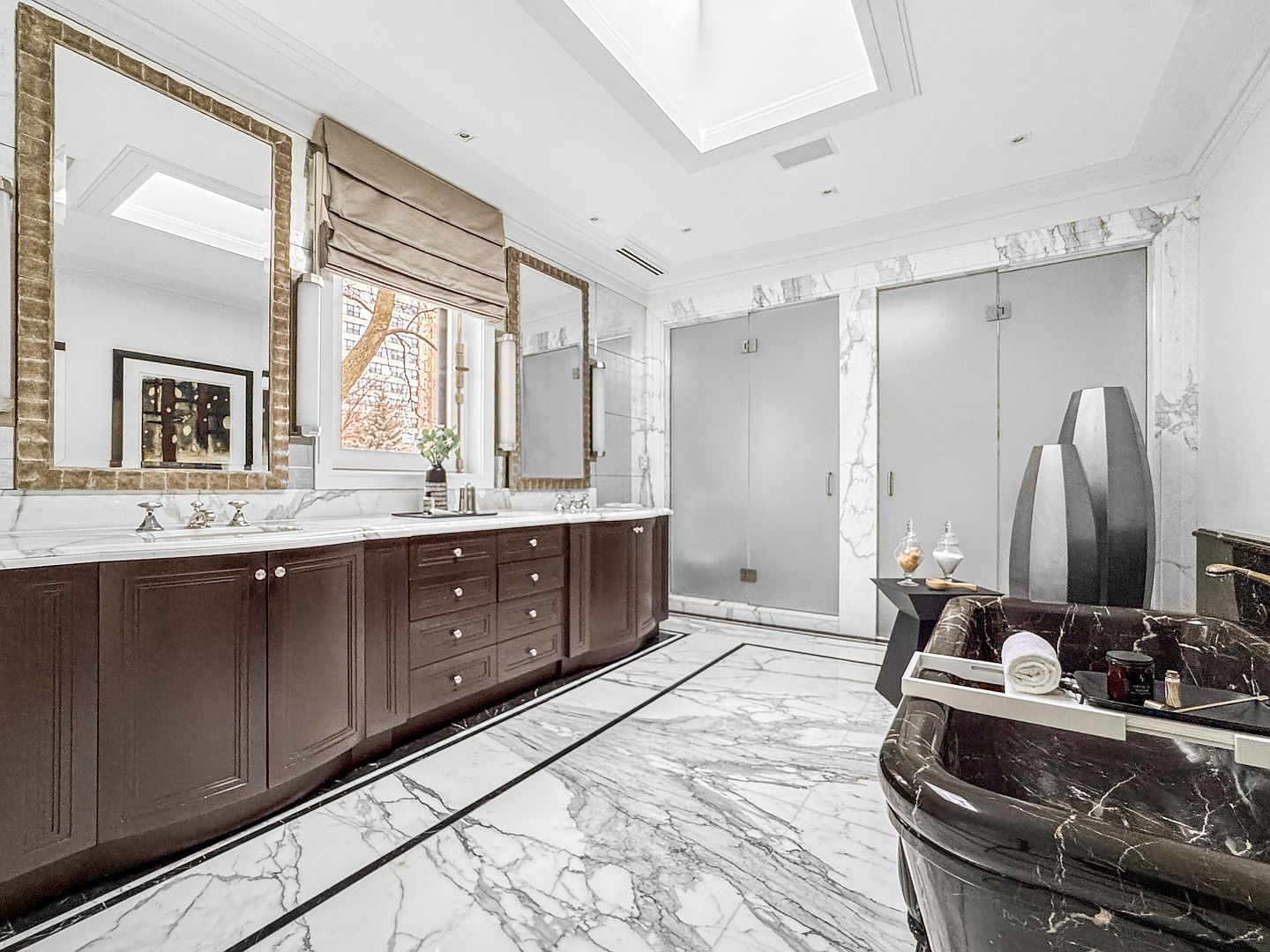 This luxurious primary bathroom features a dark-toned vanity with dual sinks, topped with light-veined stone counters and framed by large, ornate mirrors. A dark, polished stone soaking tub sits in the foreground, while the room is finished with high-contrast patterned stone flooring and frosted glass doors leading to private areas. The space is illuminated by a skylight and elegant wall sconces, creating a sophisticated and high-end atmosphere.