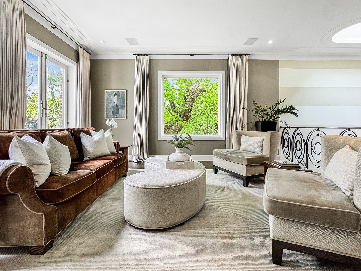 This elegant living room features a rich brown velvet sofa paired with two neutral-toned armchairs and a unique, curved ottoman centerpiece. Large windows draped in floor-length curtains frame lush green views, while a textured area rug anchors the seating arrangement. The space is accented by a framed piece of art and a decorative plant, creating a sophisticated and inviting atmosphere.