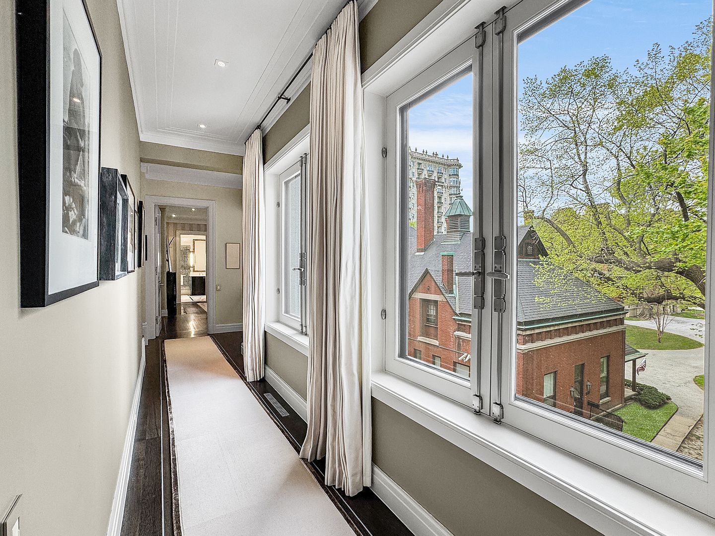 This elegant hallway features dark hardwood floors topped with a long, light-colored runner, creating a sophisticated contrast. Tall windows with cream-colored drapery line the right wall, offering a clear view of the neighboring brick architecture and lush green trees outside. The perspective looks down the corridor toward an open doorway, showcasing a bright, well-lit transition space with framed artwork adorning the left wall.