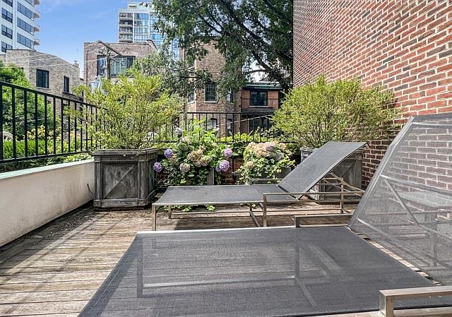 This rooftop deck features wooden plank flooring and two modern lounge chairs positioned for relaxation. The space is accented by large planters filled with lush greenery and blooming hydrangeas, set against a backdrop of a brick building and urban architecture. The perspective is a low-angle shot looking across the deck, emphasizing the private outdoor living area.