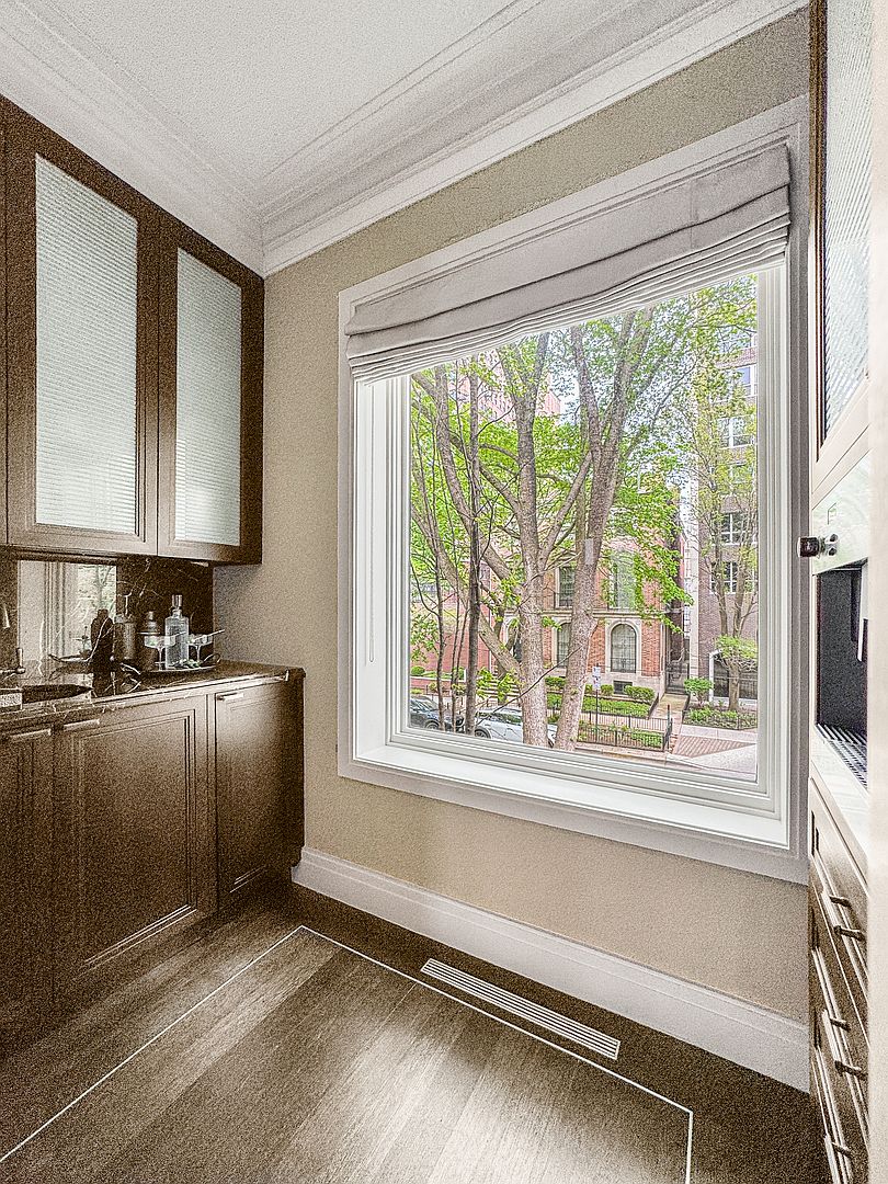 This elegant butler's pantry or wet bar area features dark wood cabinetry with frosted glass upper doors and a dark stone countertop. A large window with a neutral fabric roman shade provides a bright, scenic view of mature trees and neighboring brick architecture. The space is finished with dark wood flooring and a clean, sophisticated aesthetic suitable for entertaining.