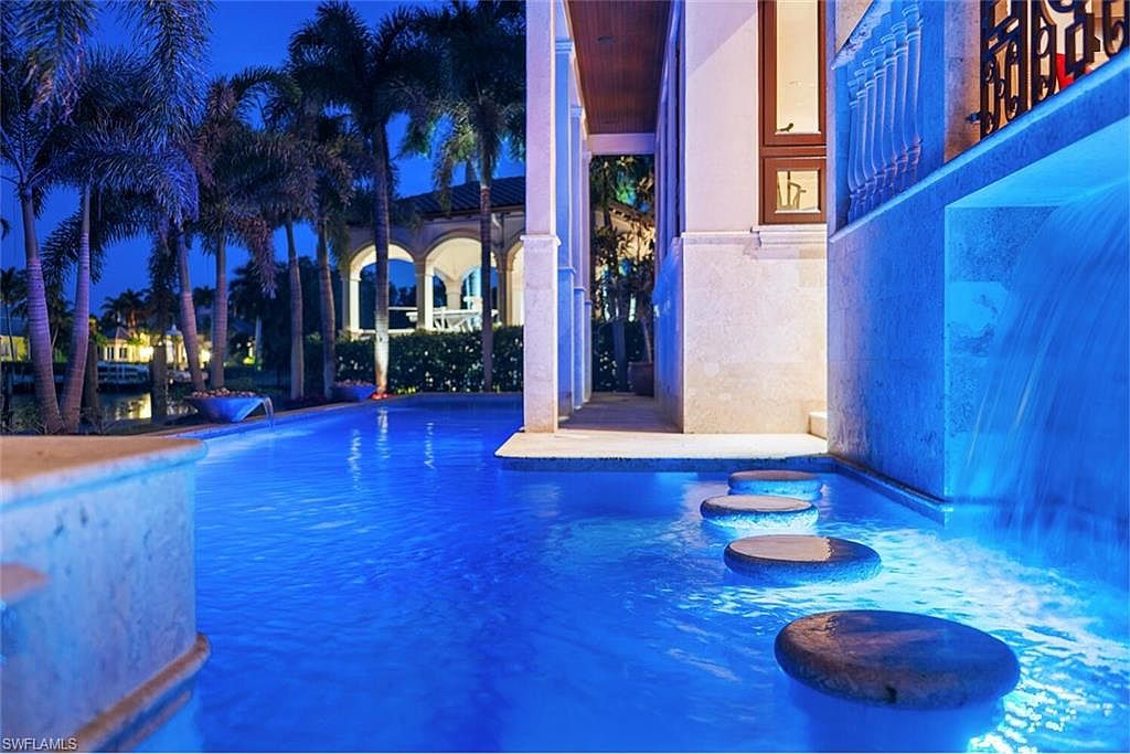 This image showcases a luxurious pool area at twilight, featuring stepping stones leading across the water and a waterfall feature integrated into the architecture. Palm trees line the background, adding to the tropical ambiance. The pool is illuminated with blue lighting, creating a serene and inviting atmosphere, perfect for relaxation and entertainment.
