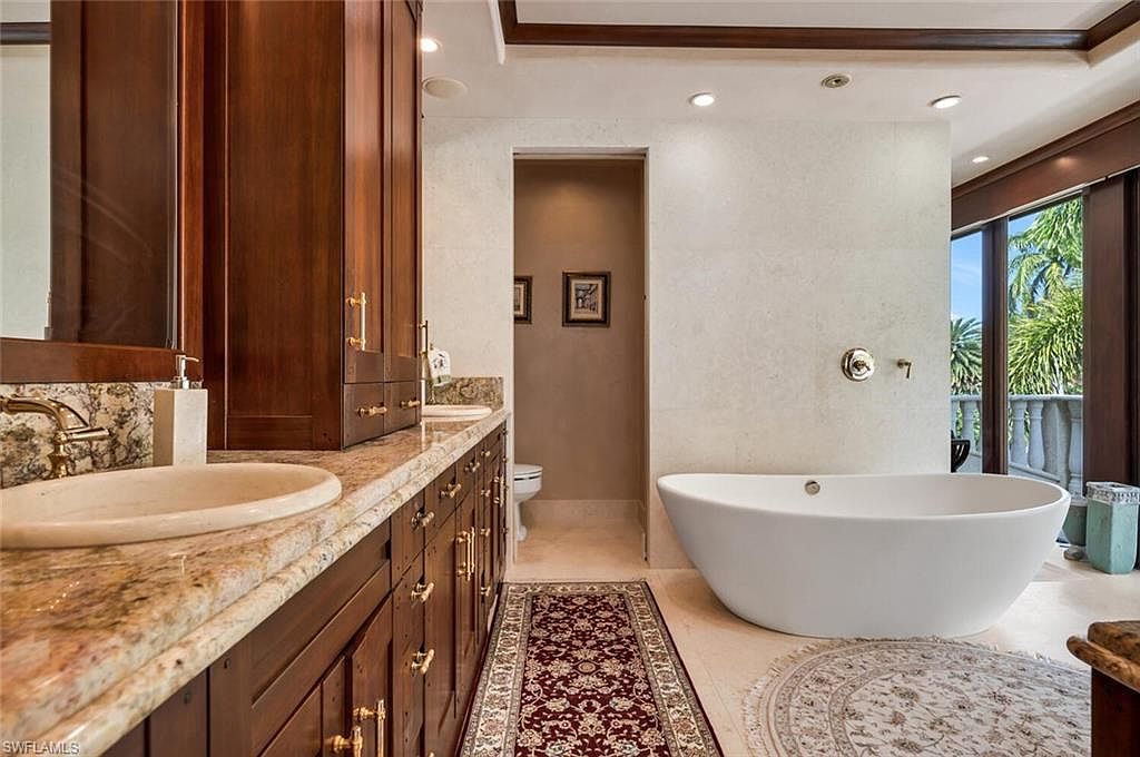 This is a luxurious primary bathroom featuring a freestanding white bathtub, a long vanity with dark wood cabinets and granite countertops, and a separate toilet room. The walls are covered in light-colored stone tiles, and the space is illuminated by recessed lighting. A large window offers a view of the outdoors, enhancing the spa-like atmosphere.