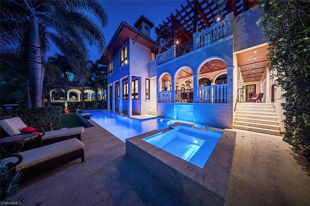 This image showcases a luxurious backyard featuring a stunning pool and spa area at night. The pool is illuminated with a vibrant blue light, creating a serene and inviting atmosphere. The architecture of the house is visible in the background, with arched balconies and lush greenery adding to the overall elegance of the outdoor space.