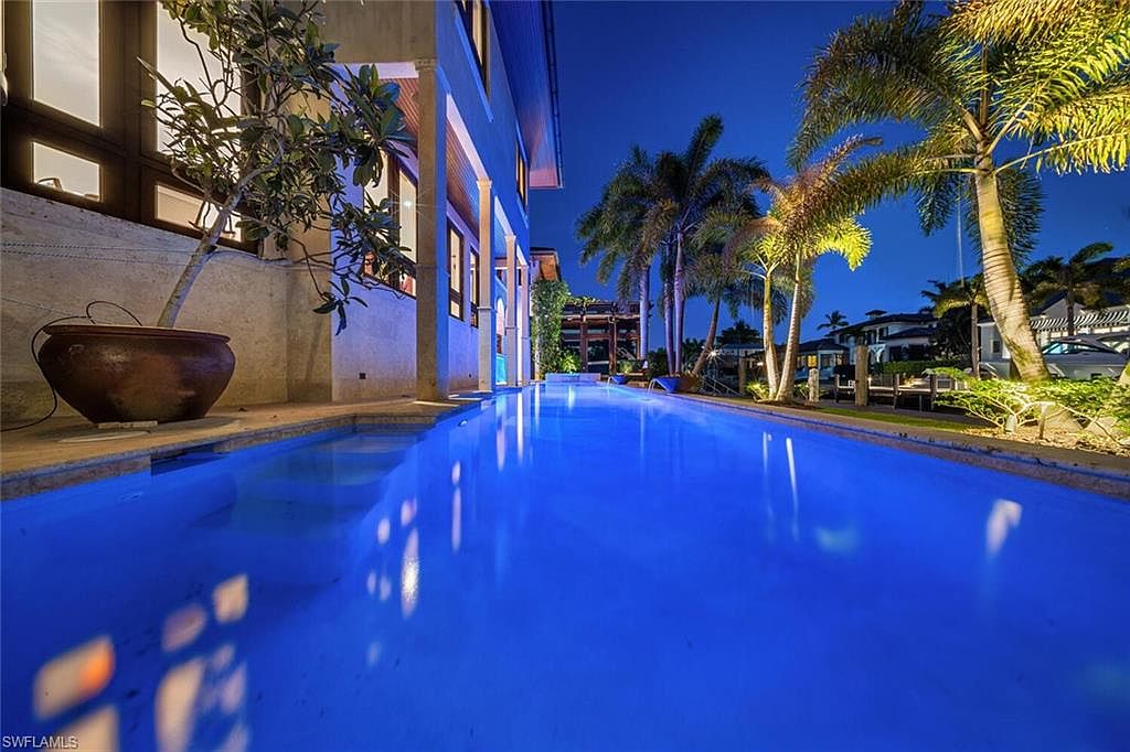 This image showcases a luxurious outdoor pool area at night, featuring a long, illuminated pool with vibrant blue water. Palm trees line the side of the pool, adding a tropical feel, while the architecture of the adjacent building suggests a high-end property. The scene evokes a sense of relaxation and upscale living.