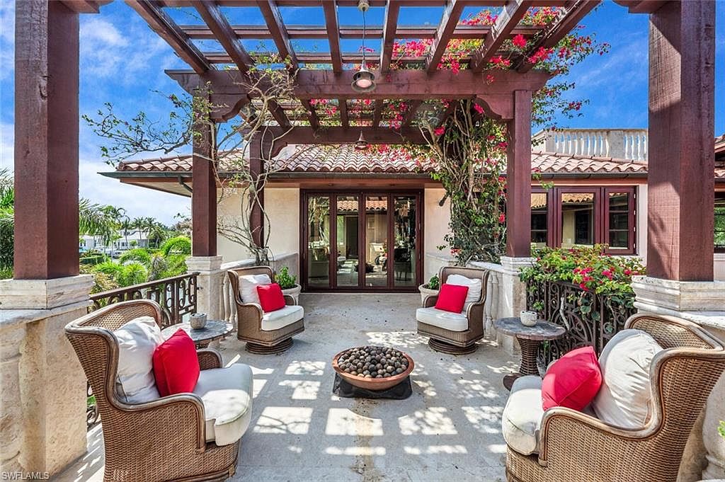 This image showcases a luxurious outdoor patio area, featuring comfortable wicker seating arranged around a central fire pit. The patio is covered by a pergola adorned with flowering vines, casting dappled sunlight across the space. The scene exudes a sense of relaxation and upscale outdoor living, perfect for entertaining or enjoying quiet moments.