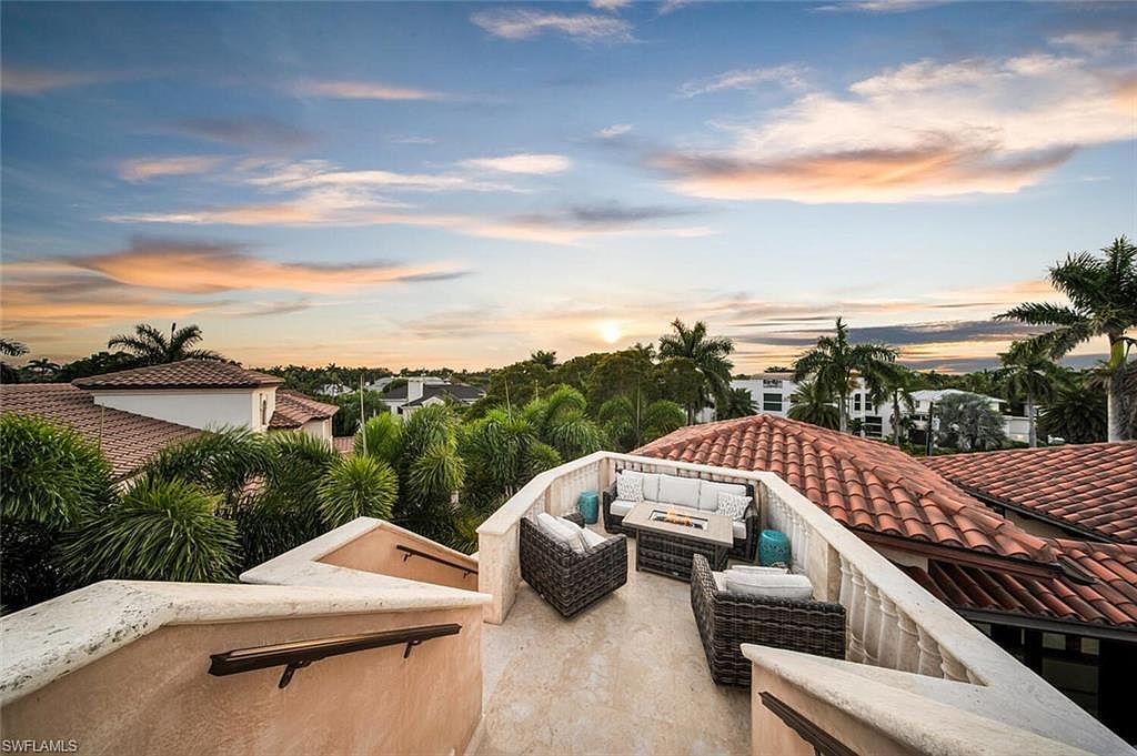 This image showcases a luxurious rooftop patio at sunset, featuring comfortable outdoor seating arranged around a modern fire pit. The patio is surrounded by a low wall with decorative railings, offering panoramic views of the surrounding neighborhood with lush greenery and tiled roofs. The overall impression is one of upscale relaxation and outdoor living.