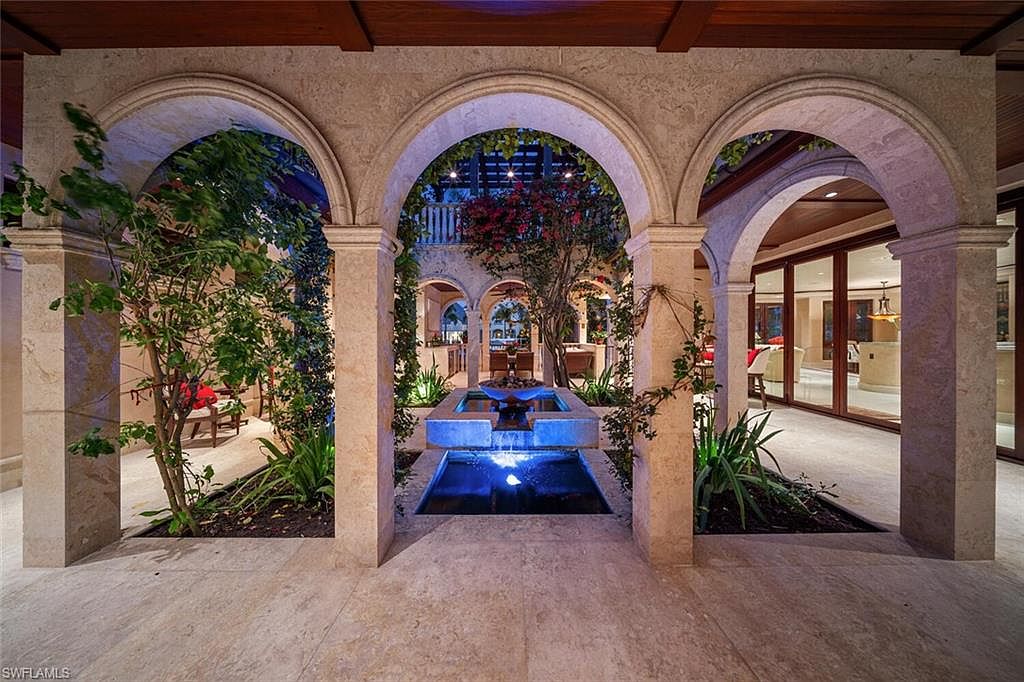 This image showcases a stunning outdoor patio area featuring a series of arched openings supported by stone columns, creating a courtyard effect. A multi-tiered fountain with blue lighting serves as a focal point, surrounded by lush greenery and manicured landscaping. The space exudes a luxurious and tranquil ambiance, perfect for relaxation and entertainment.