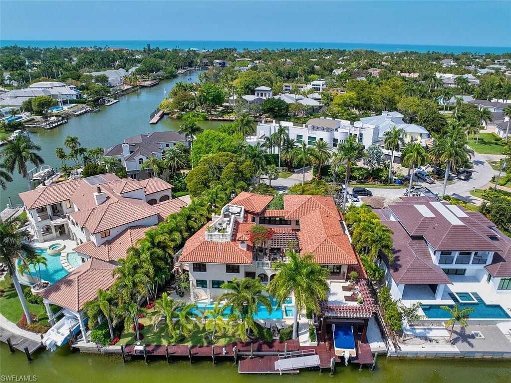This aerial shot showcases a luxurious waterfront property featuring a terracotta-tiled roof, a private pool, and a boat dock. Lush landscaping surrounds the home, enhancing its curb appeal and creating a serene atmosphere. The property is situated on a canal, offering direct access to the water and stunning views of the surrounding neighborhood.