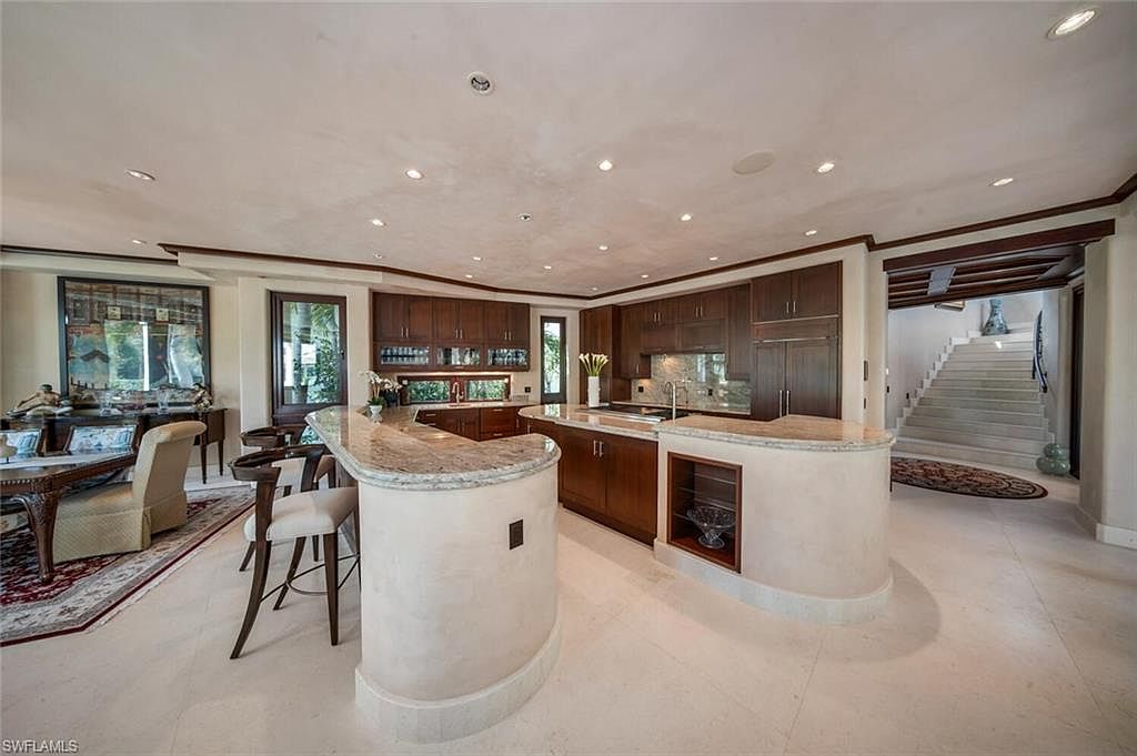 This interior shot showcases a spacious kitchen with dark wood cabinetry and a unique curved island featuring a light-colored countertop. The kitchen seamlessly transitions into a dining area and a hallway with stairs, creating an open and inviting atmosphere. The flooring is a light tile, enhancing the brightness of the space.