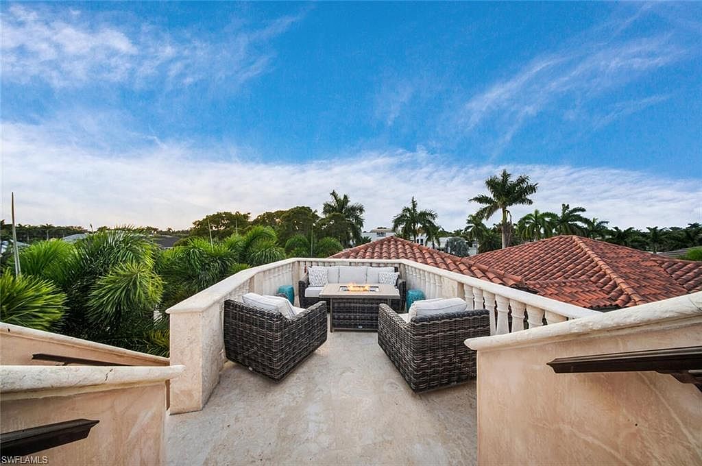 This image showcases a stylish rooftop patio or balcony area, furnished with a modern outdoor seating arrangement including a sofa, chairs, and a fire pit table. The space is surrounded by a low wall with decorative balusters, offering views of lush greenery and tiled rooftops in the distance under a bright blue sky. The overall impression is one of luxury and relaxation.