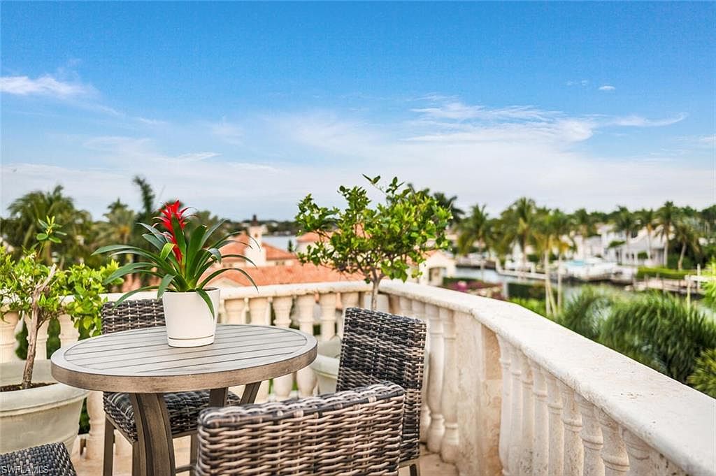 This image showcases a charming balcony or patio area with a round table and wicker chairs, creating an inviting outdoor living space. The balcony features a decorative stone railing, and the view includes lush greenery, palm trees, and waterfront homes, suggesting a tropical or coastal setting. The scene evokes a sense of relaxation and luxury.