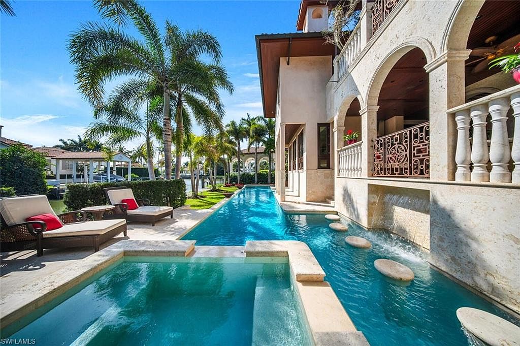 This image showcases a luxurious outdoor pool and spa area of a high-end property. The pool features a unique design with stepping stones and a waterfall feature, complemented by lounge chairs for relaxation. Lush landscaping and palm trees enhance the tropical ambiance, creating an inviting and serene outdoor living space.