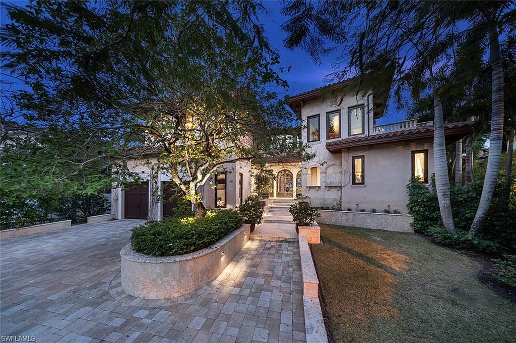 This is a front exterior view of a luxurious two-story home with a Mediterranean architectural style. The house features a light-colored stucco facade, a red tile roof, and multiple windows with dark frames. A paved driveway leads to a two-car garage on the left, while a landscaped walkway with stone pavers and illuminated garden beds guides visitors to the arched entryway. Mature trees and manicured lawns enhance the property's curb appeal, creating an inviting and upscale impression.