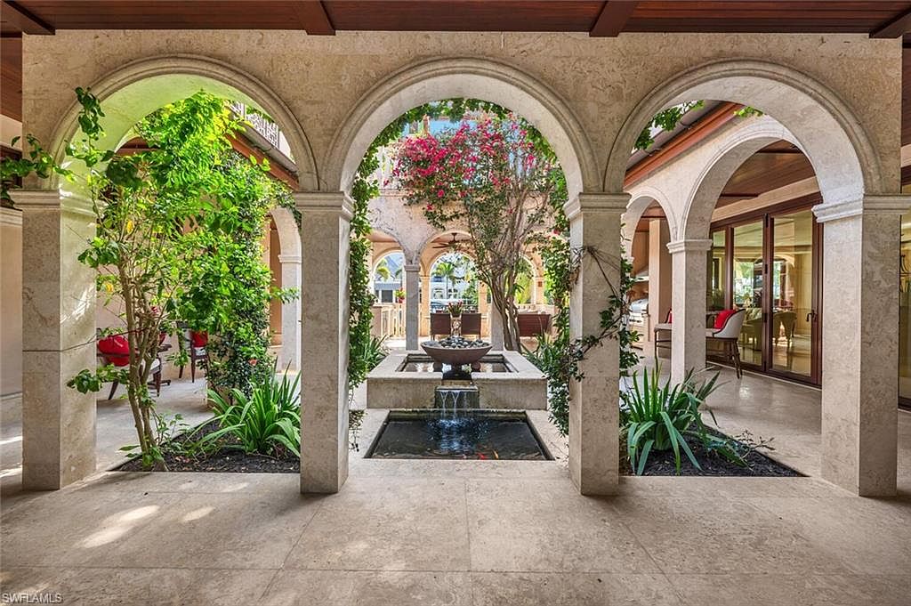 This image showcases a stunning outdoor patio area with a series of arched openings supported by stone columns, creating a courtyard effect. Lush greenery and a central water feature add to the serene ambiance. The patio is paved with light-colored stone, and glimpses of outdoor seating and interior spaces can be seen through the arches, suggesting a seamless indoor-outdoor living experience.