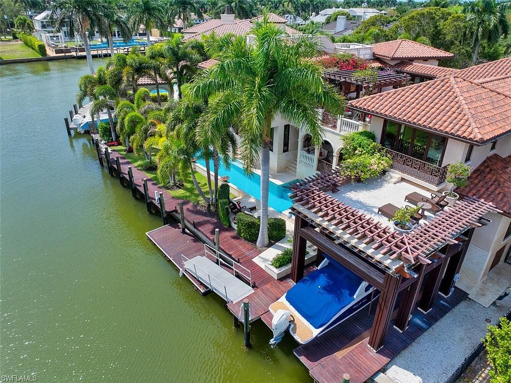 This aerial view showcases a luxurious waterfront property with a Mediterranean-style house featuring a terracotta tile roof, multiple balconies, and a private pool. The property includes a boat dock with a boat lift and several palm trees lining the canal, enhancing the tropical ambiance. The overall impression is one of upscale coastal living.