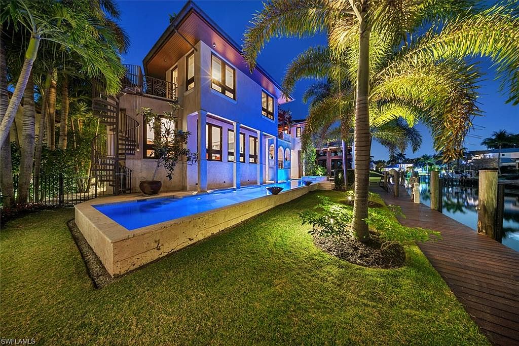 This exterior shot showcases a luxurious home with a stunning pool that extends along the side of the house, leading to a waterfront dock. The property features lush landscaping with palm trees, and the architectural design includes a spiral staircase and multiple windows, creating an inviting and upscale ambiance. The image captures the essence of waterfront living with a blend of modern design and tropical charm.