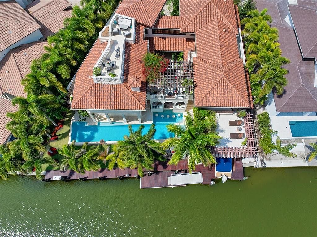This aerial shot showcases a luxurious waterfront property with a terracotta tile roof, a large swimming pool, and a private dock. Lush tropical landscaping surrounds the house, enhancing its appeal. The overall impression is one of opulence and tranquility, perfect for a high-end real estate listing.