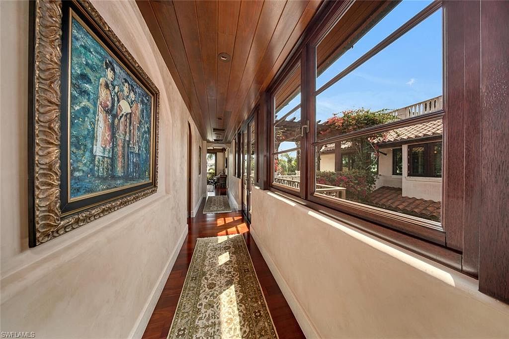 This interior shot showcases a hallway with rich wooden floors and a matching wooden ceiling, creating a warm and inviting atmosphere. A large framed painting adorns the wall, while a long rug stretches down the hallway, leading the eye towards a distant room. Large windows on the right offer views of the exterior, filling the space with natural light.