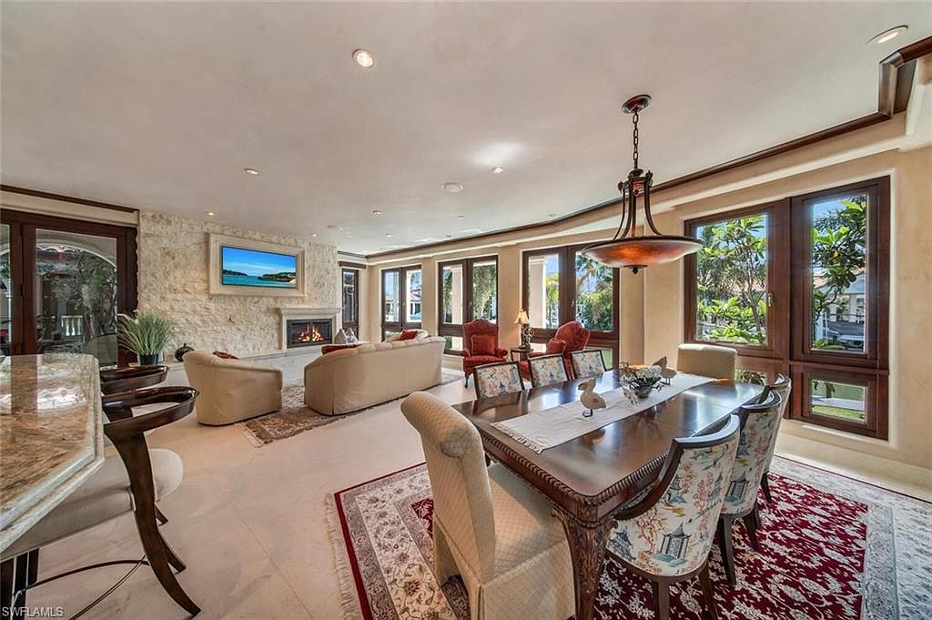 This interior shot showcases a luxurious living and dining area, featuring a stone accent wall with a fireplace and a mounted television. The space is well-lit with natural light streaming through large windows, complemented by an elegant chandelier above the dining table. The room is furnished with comfortable seating and stylish rugs, creating a warm and inviting atmosphere.