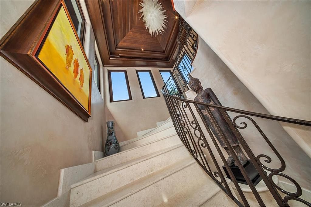 This interior shot showcases a grand staircase with marble steps and an ornate wrought iron railing featuring a statue. A painting hangs on the wall above a decorative vase, while natural light streams in through a series of windows. The ceiling is wood paneled with a modern light fixture, creating a luxurious and elegant atmosphere.