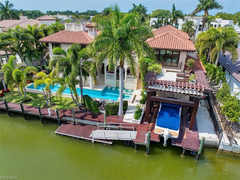 This aerial view showcases a luxurious waterfront property with a Mediterranean-style home featuring a terracotta tile roof, a private pool, and a boat dock with a boat lift. Lush tropical landscaping surrounds the property, enhancing its appeal and privacy. The overall impression is one of upscale coastal living.