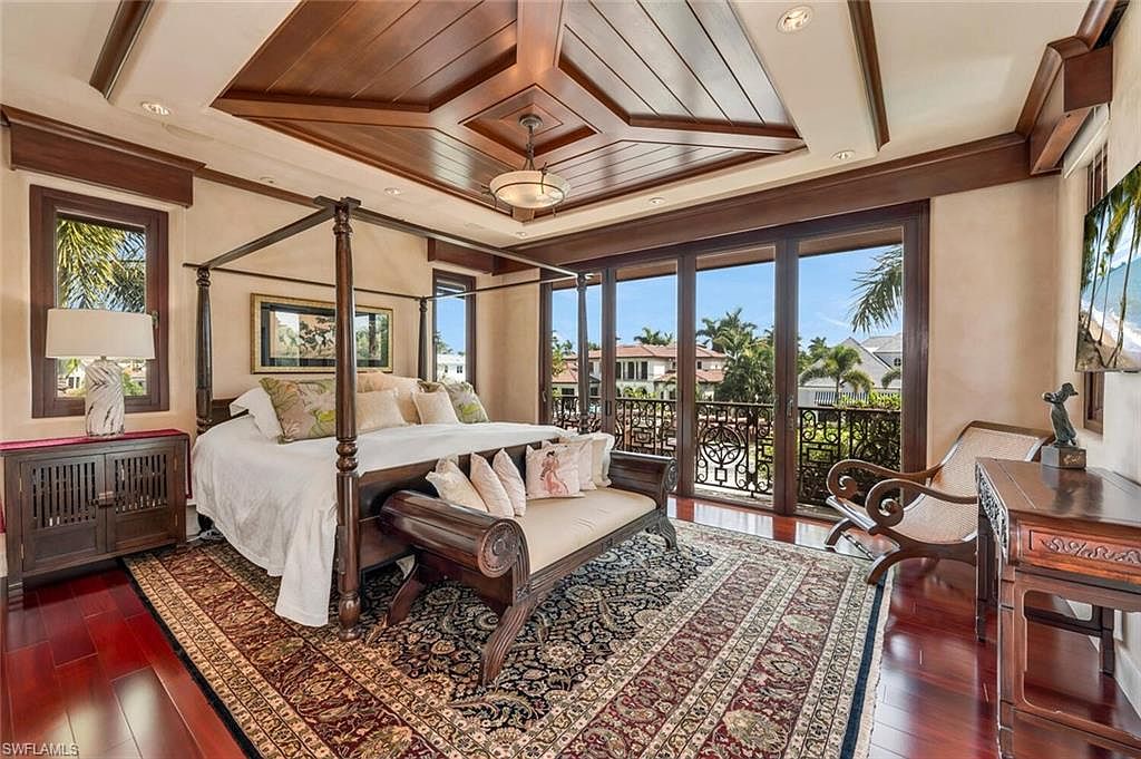 This is a luxurious primary bedroom featuring a four-poster bed with elegant bedding and a bench at the foot. The room boasts a detailed wood ceiling, a large area rug, and sliding glass doors leading to a balcony with views of the surrounding area. The overall impression is one of sophistication and comfort.