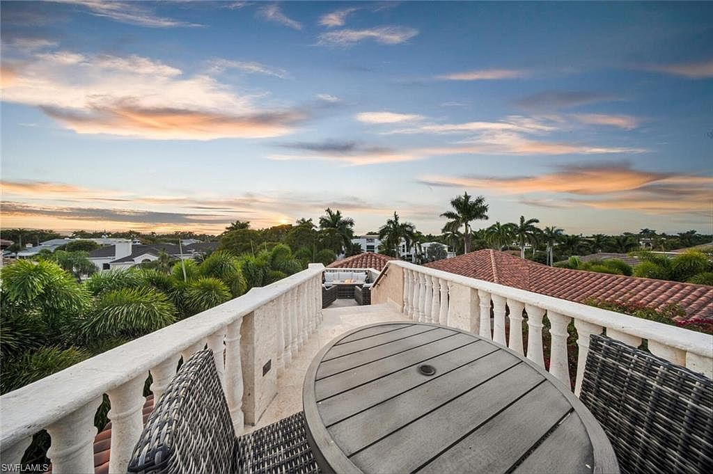 This image showcases a luxurious rooftop balcony with a stunning view. The balcony features a round wooden table and wicker chairs, perfect for outdoor dining or relaxation. A decorative balustrade surrounds the perimeter, and in the background, lush greenery, palm trees, and glimpses of nearby houses can be seen under a beautiful sunset sky.