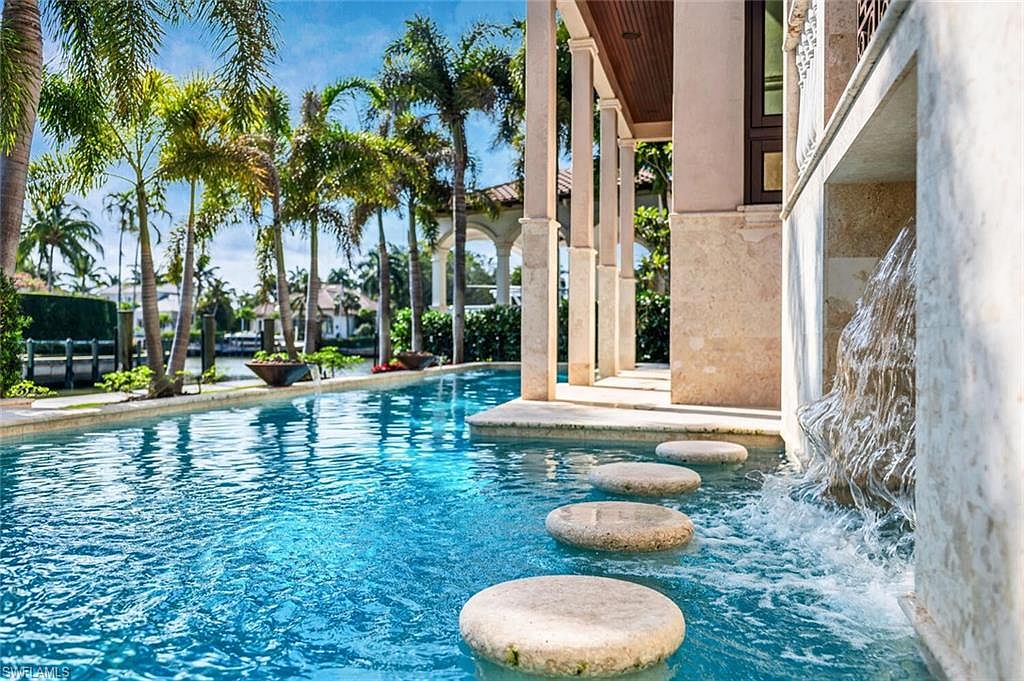 This image showcases a luxurious pool area with stepping stones leading across the water. The pool is surrounded by lush palm trees and elegant architectural features, including columns and a waterfall feature. The scene evokes a sense of tranquility and upscale living, perfect for attracting potential buyers seeking a resort-like experience at home.