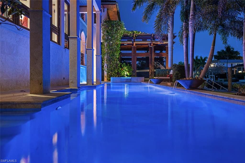 This image showcases a luxurious outdoor pool area at night, featuring illuminated water and elegant architectural details. The pool is surrounded by columns and lush greenery, with water features adding to the ambiance. Palm trees frame the scene, creating a tropical and upscale atmosphere, perfect for relaxation and entertainment.