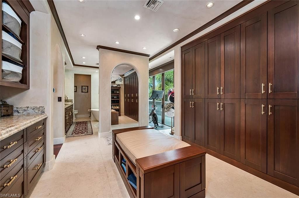 This is a well-organized walk-in closet featuring dark wood cabinetry and a central bench with storage. The closet is spacious, with ample storage for clothing and accessories. The design incorporates arched doorways leading to other areas, suggesting a luxurious and functional space.
