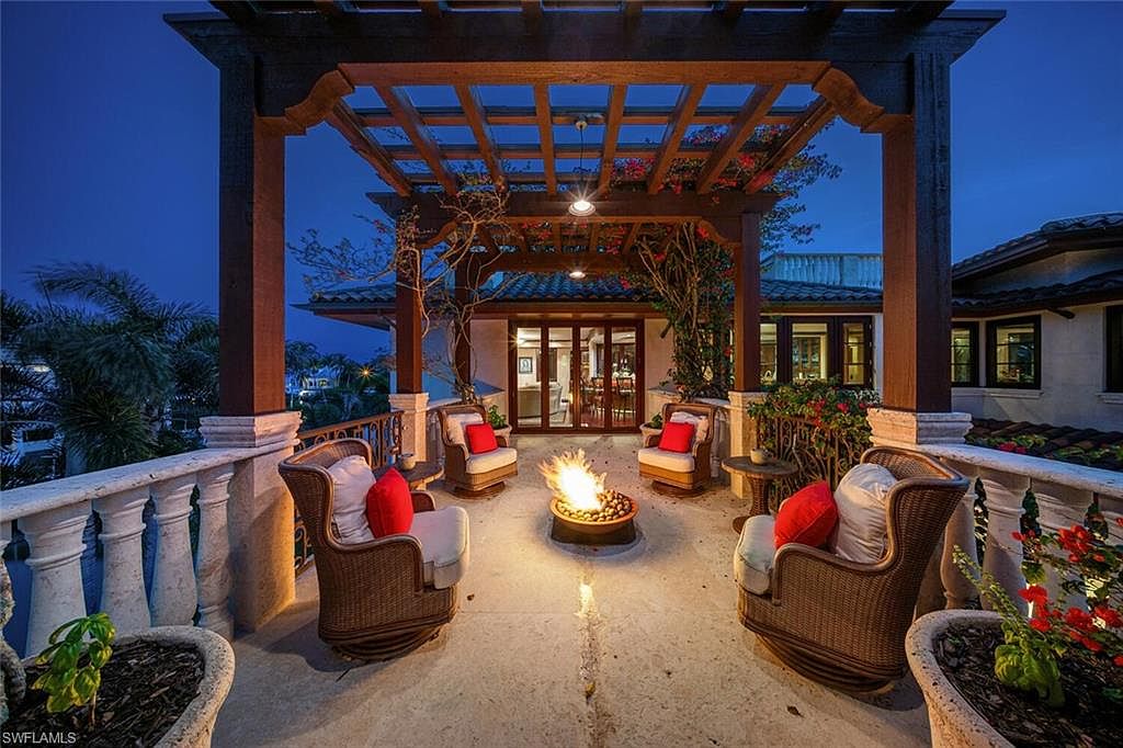 This image showcases a luxurious outdoor patio area at dusk, featuring a pergola-covered seating arrangement with four wicker chairs adorned with plush cushions and red pillows. A central fire pit adds warmth and ambiance, while the surrounding balustrade and potted plants enhance the elegant atmosphere. The view extends beyond the patio, hinting at a well-maintained property and serene surroundings.