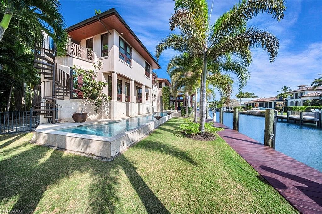 This image showcases the rear exterior of a luxurious waterfront home, featuring a modern architectural design with a long, narrow pool adjacent to the building. Palm trees and manicured green lawn lead to a private dock on the canal, creating a serene and upscale ambiance. The spiral staircase adds a unique architectural element.