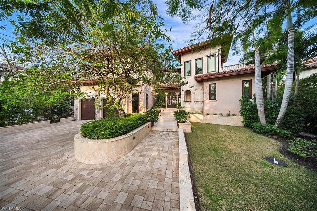 This is a front view of a luxurious Mediterranean-style home. The property features a stone driveway and walkway leading to the arched entryway. Lush landscaping, including mature trees and manicured lawns, surrounds the house, enhancing its curb appeal and creating a welcoming atmosphere.